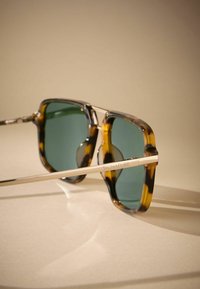 Sunglasses with a tortoiseshell frame, green lenses, and a sleek metal arm featuring the word "SIGNATURE" engraved.
