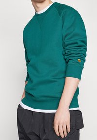 Sudadera turquesa con cuello redondo, puños y dobladillo acanalados. Presenta un pequeño logotipo naranja en el puño. Llevada sobre una camiseta blanca.