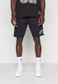 Bermudas de algodão pretas com logótipo, combinadas com meias brancas até ao tornozelo e tênis brancos. Design simples, corte padrão, bordas limpas.
