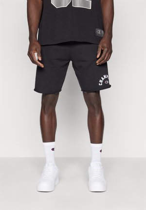 Pantalones cortos de algodón negros con logo, combinados con calcetas blancas y zapatillas blancas. Diseño sencillo, ajuste estándar, bordes limpios.