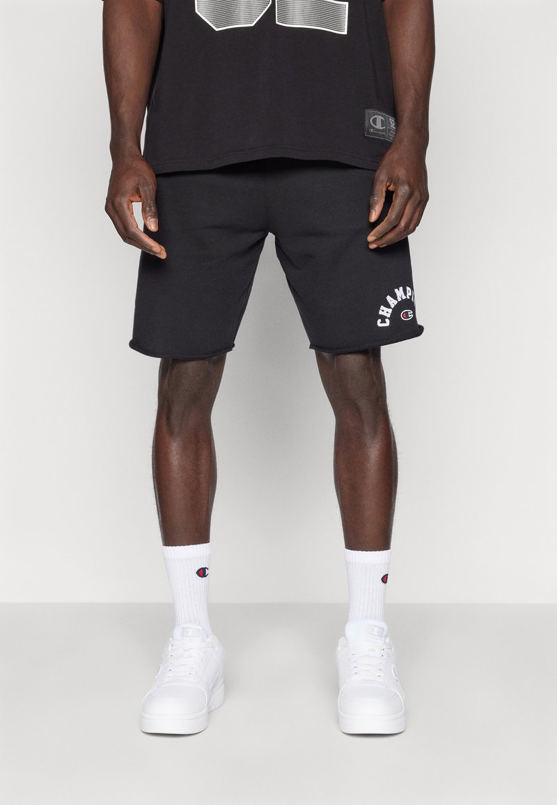 Bermudas de algodão pretas com logótipo, combinadas com meias brancas até ao tornozelo e tênis brancos. Design simples, corte padrão, bordas limpas.