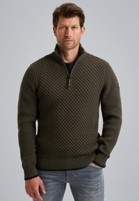Olijfgroen gebreid sweater met een gestructureerd ruitpatroon, halve ritskraag en geribbelde manchetten, gecombineerd met lichtgrijze jeans.