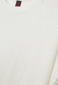 Weißes Baumwoll-T-Shirt mit weicher Textur, ausgestattet mit einem Rundhalsausschnitt und einem dezenten melierten Muster. Etikett sichtbar mit roten und schwarzen Farben.