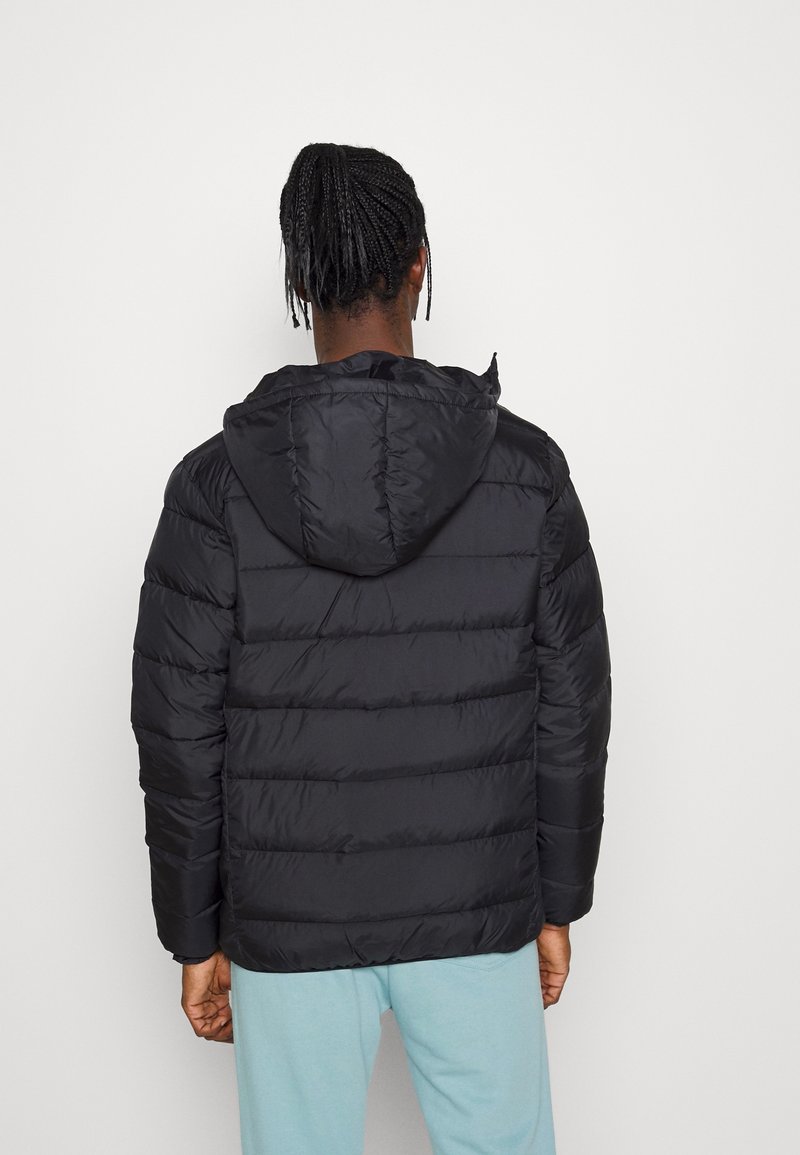 Personne aux cheveux tressés vue de dos, portant une veste à capuche matelassée noire et un pantalon bleu clair sur un fond blanc uni.