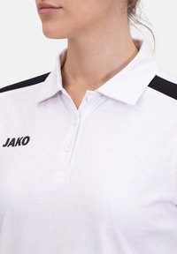 Polo bianco con colletto a punta, con un design nero sulla spalla e la parola "JAKO" in nero sul lato sinistro.