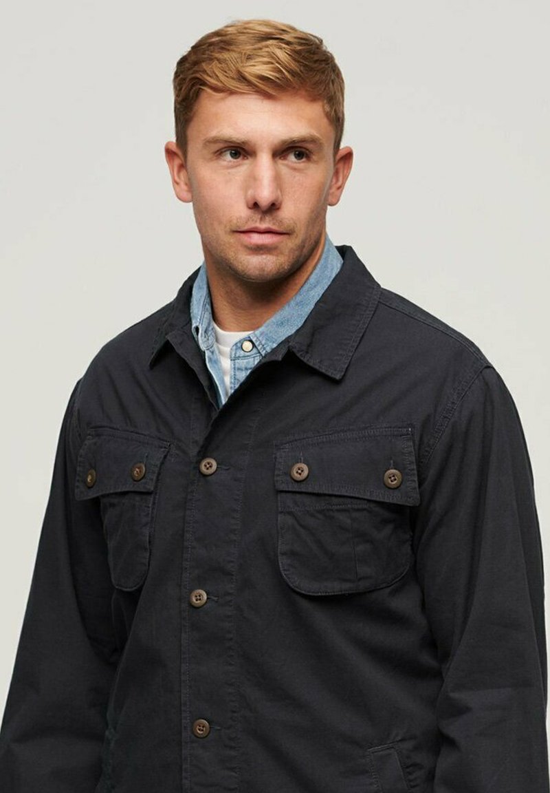 Chaqueta de un oscuro color navy hecha de algodón, con dos bolsillos en el pecho con botones marrones, diseño de cuello y un acabado de tejido texturizado.