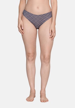 Frau steht barfuß und trägt blau-beige karierte Bikinihose vor einem schlichten hellen Hintergrund.