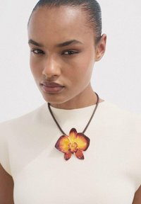 Bruine leren ketting met een grote, levendige oranje en gele orchidee hanger. Model draagt een aansluitend crèmekleurig topje, minimalistische styling.