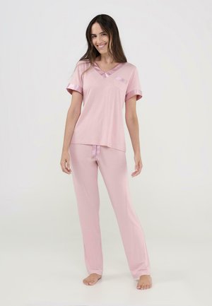 Mujer sonriendo, de pie descalza, con una blusa de manga corta rosa claro con ribete de satén y pantalones sueltos a juego, frente a un fondo claro y sencillo.