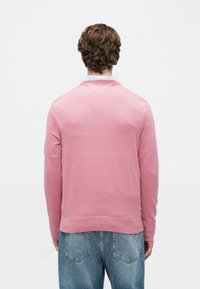 Mężczyzna z krótkimi kręconymi włosami, ubrany w różowy sweter na jasnoniebieską koszulę z kołnierzykiem i niebieskie dżinsy, stoi przodem do białego tła.