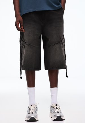 Muž v čiernych cargo kraťasoch, bielych športových ponožkách a sivobielych športových topánkach stojaci na bielom pozadí.