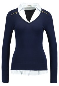Marineblauwe lange mouw trui met V-hals en een witte kraagshirt eronder. Voorzien van gouden ritsen op de schouders en geribde manchetten.
