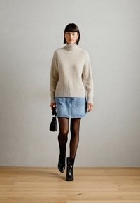 Beige gebreide coltrui, denim minirok, zwarte enkellaarsjes en een kleine zwarte handtas, staande op houten vloeren tegen een effen muur.