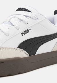 Bílá kožená teniska s černými a šedými prvky, texturovaným semišovým panelem a bílými tkaničkami. Na boku má černé logo a hnědou podrážku.