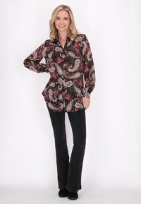 Camicia nera a maniche lunghe con stampa paisley e floreale in rosso e bianco, con chiusura a bottoni e orlo curvo. Indossata con pantaloni neri.