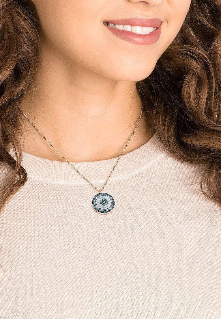 Silberne Halskette mit einem runden Anhänger, der ein blaues und filigranes Muster zeigt, an einer dünnen Kette hängend, getragen über einem hellbeigen Oberteil.