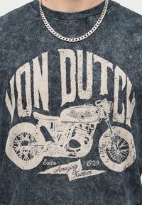 Homme portant une chemise sombre avec un grand motif vintage de moto et le texte "Von Dutch", "Since 1929" et "Amazing Kustom", plus une chaîne en argent.