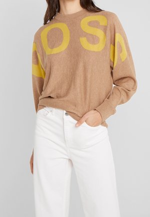 Suéter de punto marrón con grandes letras amarillas "S" y "A" en las mangas, escote redondo y corte relajado. Combinado con jeans blancos.