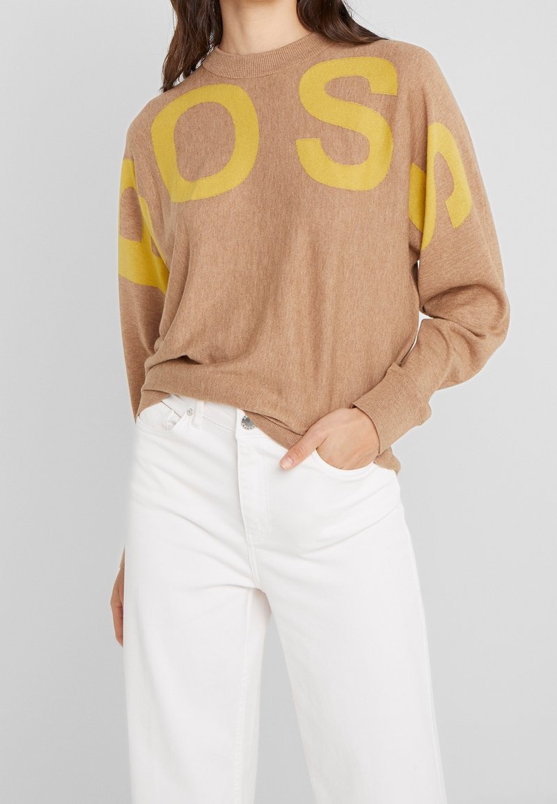 Brauner Strickpullover mit großen gelben Buchstaben "S" und "A" auf den Ärmeln, Rundhalsausschnitt und lockerer Passform. Kombiniert mit weißen Jeans.