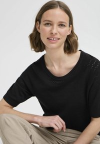 Zwarte gebreide sweater met korte mouwen, voorzien van een textuurpatroon en een ronde halslijn, gecombineerd met lichtbeige broeken.