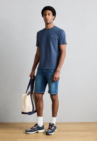 Granatowy bawełniany T-shirt z krótkimi rękawami i małym logo, w połączeniu z dżinsowymi szortami, białymi skarpetkami i wielokolorowymi trampkami, trzymając torbę typu tote.