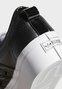 Černé perforované kožené tenisky s lesklým patním tabem a bílou gumovou podrážkou. Obsahuje značkovou nálepku na boku.