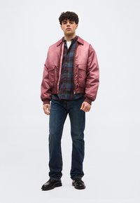 Νέος άνδρας στέκεται φορώντας ροζ μπουφάν bomber, μπλε πουκάμισο με καρό σχέδιο, σκούρο μπλε τζιν και μαύρα παπούτσια μπροστά σε μονόχρωμο γκρι φόντο.