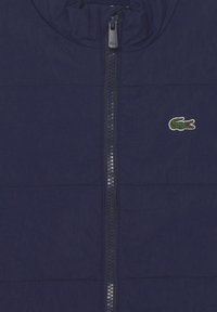 Gilet trapuntato blu navy con collo alto, zip a tutta lunghezza e un piccolo logo in ricamo a forma di coccodrillo sul lato sinistro del petto. Tessuto testurizzato.