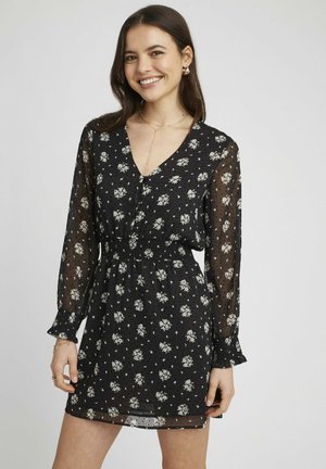 Robe noire florale en tissu léger et transparent, avec des manches longues, un décolleté en V et une taille élastique ornée de petites fleurs blanches.