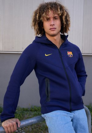 Hoodie bleu marine avec une fermeture éclair sur toute la longueur à l'avant, logo jaune de Nike et blason du FC Barcelone, comprenant deux poches latérales zippées et une capuche doublée.