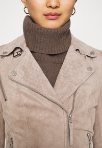 Femme portant une veste en daim beige perforée avec des fermetures éclair argentées, par-dessus un pull à col roulé côtelé marron et de petites boucles d'oreilles créoles dorées.
