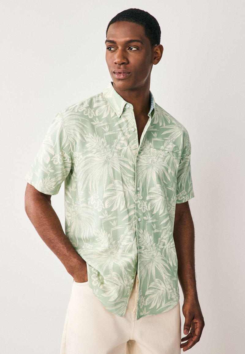 Camicia a maniche corte con bottoni in verde chiaro con motivo di foglie tropicali bianche, caratterizzata da un colletto classico e una tasca sul petto sinistro.
