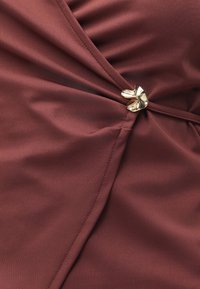 Κοντινή λήψη από μπορντό ύφασμα μαζεμένο και στερεωμένο με μια μικρή γυαλιστερή χρυσή αγκράφα σε αφηρημένο σχήμα.