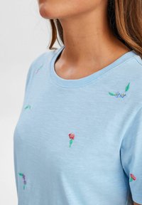 Camisa de manga corta azul claro con diseños florales bordados en rosa, morado y verde. Textura suave con cuello redondo.