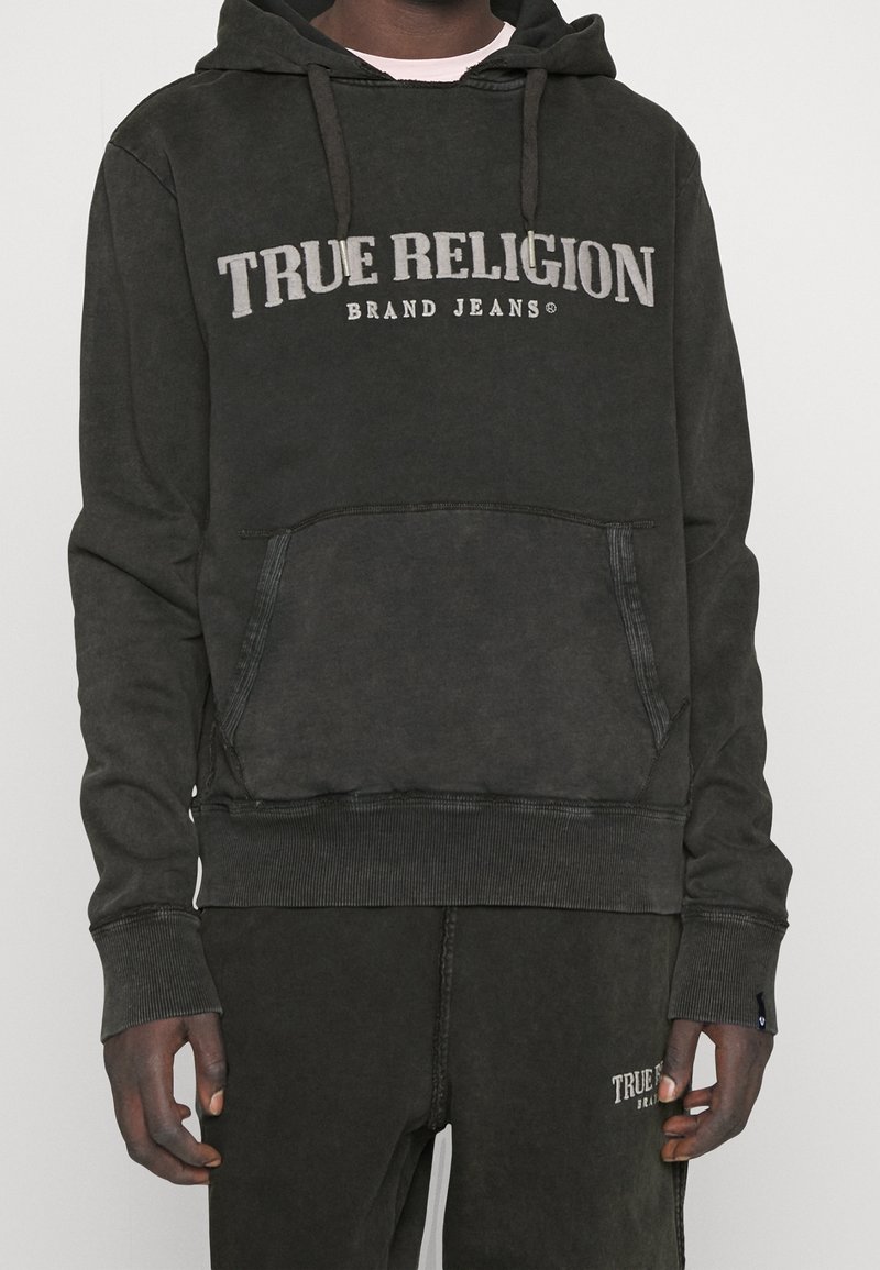 Sweat à capuche gris foncé avec une poche avant, des poignets et un ourlet côtelés. Présente le texte "TRUE RELIGION BRAND JEANS" en gris clair sur la poitrine.