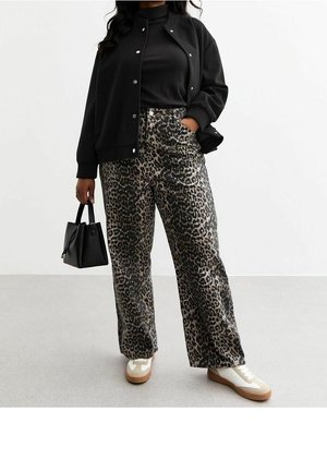 Person iført sort jakke, sort rullekrave, leopardprint bukser, hvide sneakers, holder en sort håndtaske, stående på en grå gulv.