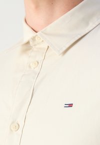 Camicia button-up beige con colletto a punta e una trama sottile. Presenta un piccolo logo ricamato in blu, rosso e bianco sul lato sinistro del petto.