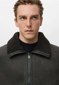 Giacca zip nera scura con colletto in shearling nero testurizzato, presentata su uno sfondo uniforme, con etichetta visibile sull zipper.