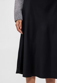 Sort satin nederdel med en glat tekstur, der har et flydende design og en nederste kant, der blødt buer. Grå sweaterærme synlig.