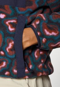Fleece-Jacke mit einem mehrfarbigen abstrakten Muster in den Farbtönen Braun, Pink und Türkis. Details sind ein marineblauer Stoffbesatz und ein verstellbarer Kordelzug.