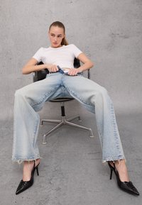Light blue, wide-leg denim jeans with frayed hems. Paired with sleek black pointed-toe heels. Model seated on a gray chair against a concrete backdrop.