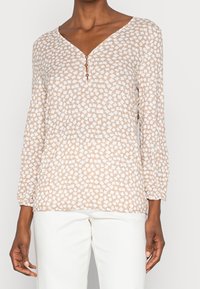 Maglia beige a maniche lunghe con scollo a V e motivo geometrico bianco, dotata di una pattina con tre bottoni; tessuto leggero e strutturato.
