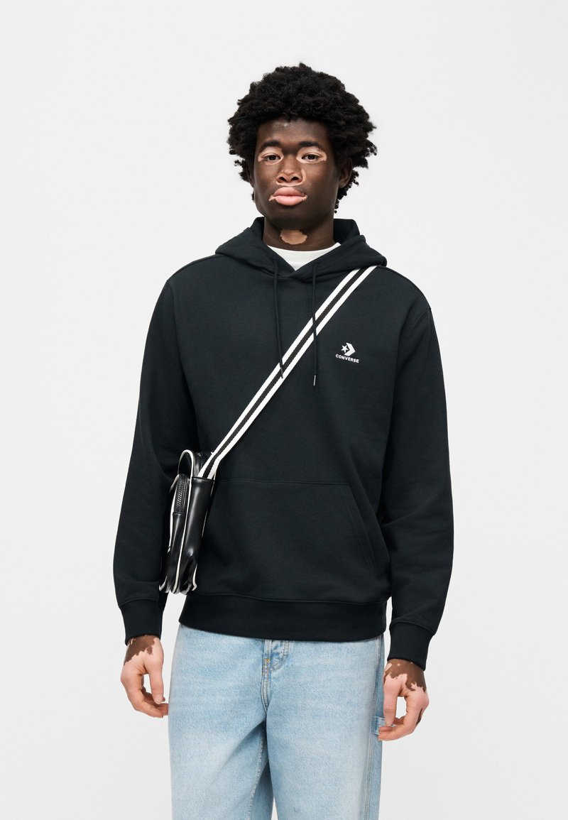 Sweat à capuche en coton noir avec logo blanc, doté d'une poche kangourou et d'une capuche à cordon ajustable. Porté avec un jean en denim bleu clair et un sac bandoulière.