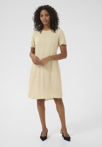 Abito beige fino al ginocchio con maniche corte, collo rotondo, due tasche laterali e texture liscia, abbinato a scarpe col tacco nere.