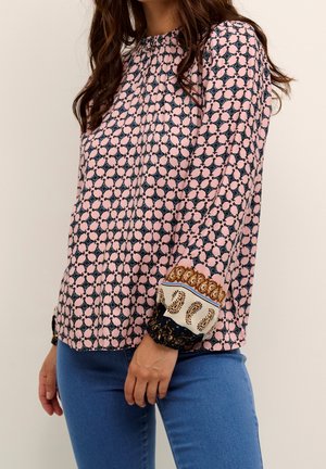 Femme portant un chemisier rose à manches longues avec un motif géométrique noir et des poignets décoratifs, associé à un jean bleu sur un fond uni.