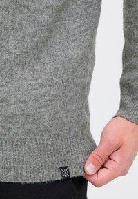 Harmaa villa neule, jossa on raidoitettu helma, ja käsi, joka pitää alaosasta kiinni. Mukana pieni musta etiketti, jossa on logo.