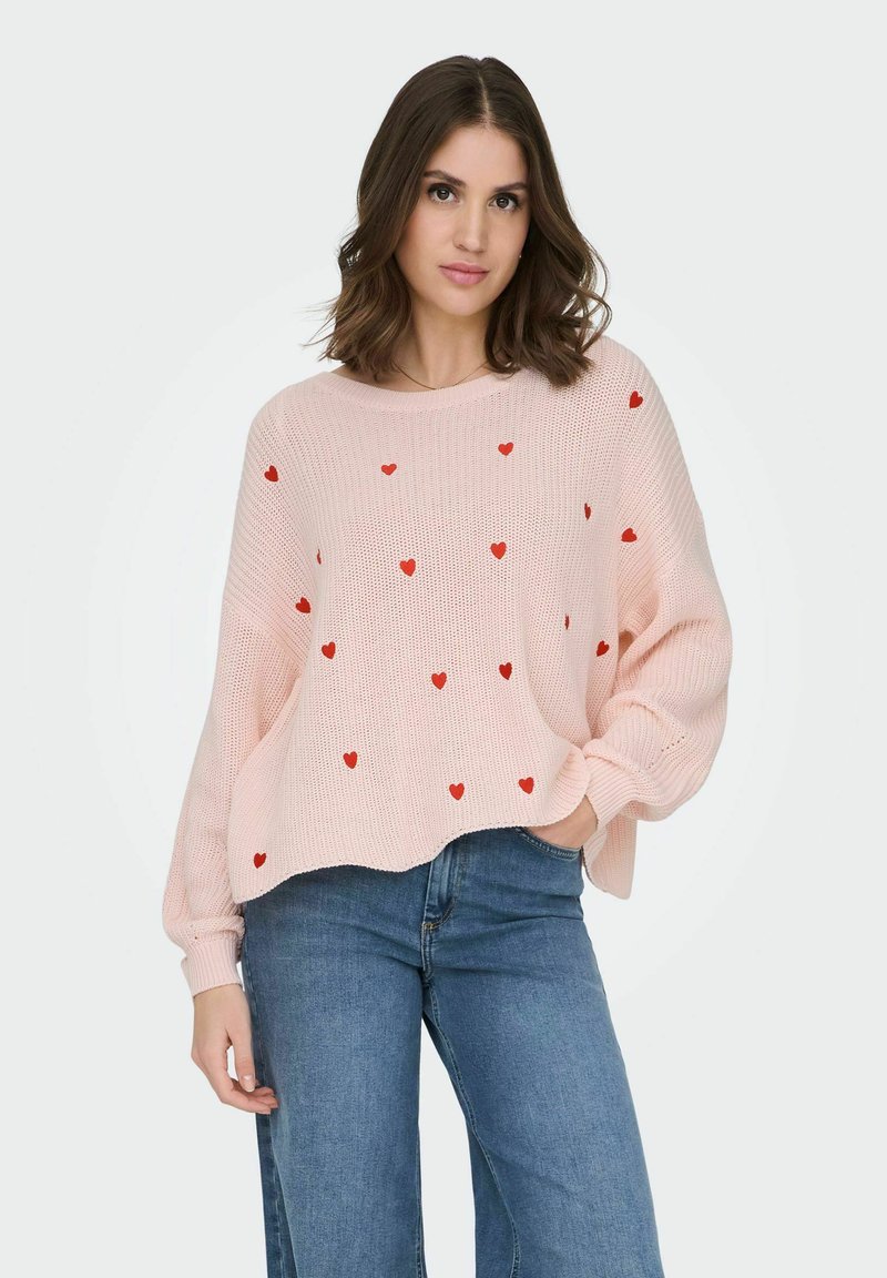 Femme portant un pull léger rose clair en tricot avec de petits cœurs rouges et un jean bleu, debout avec une main dans la poche devant un fond uni.