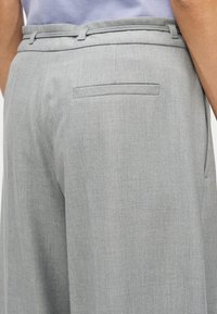 Nærbillede af en person iført lysegrå skræddersyede bukser med en baglomme med paspolering og bæltestropper, kombineret med en lys lilla top.