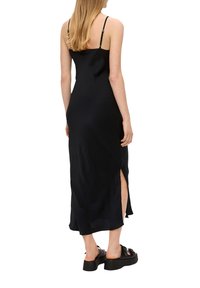 Schwarzes Maxikleid mit dünnen Trägern, seitlichen Schlitzen und glatter Textur. Getragen mit klobigen schwarzen Plateau-Sandalen.