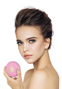 Een roze siliconen gezichtsreinigingsapparaat met textuur en gouden accenten, vastgehouden door een persoon met een heldere huid en lichte make-up.